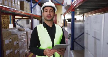 Güvenlik yeleği ve başlık takmış gülümseyen Latin lojistik teknisyeninin portresi dijital tablet deposunda duruyor. Etrafı raflardaki organize kutular ile çevrili.