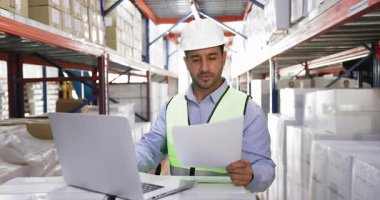 Depoda güvenlik yeleği ve miğfer giyen genç İspanyol depo çalışanı dizüstü bilgisayarla çalışıyor, kağıt belgelerini inceliyor, rapor hazırlıyor, envanteri kontrol ediyor, lojistik işlemleriyle uğraşıyor.