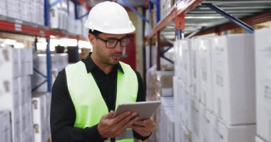 Dijital tablet kullanan, envanter ve lojistik verilerini kontrol eden, sanayi deposunda duran, güvenlik kaskı ve yelek giyen İspanyol erkek işçi. Modern depo yönetimi, tedarik zinciri işlemleri