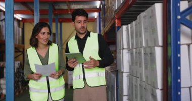 İki Latin dağıtım merkezi erkeği, tablet kullanan kadın işçiler, depo koridorunda raflara istiflenmiş karton kutularla dolu belgeleri ve envanteri gözden geçiriyorlar. Stoklama, lojistik