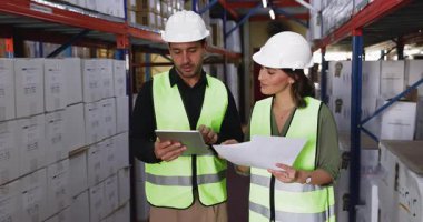 Dağıtım merkezinde çalışan Latin erkek ve kadın lojistik ekibi, tablet kullanan güvenlik yelekleri ve başlıklar giyiyorlar, belgeleri tutuyorlar, envanter verilerini tartışıyorlar, raflarda kutularla çevrili duruyorlar.