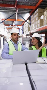 Koruyucu yelek ve şapka giyen iki genç Latin iş arkadaşı, dizüstü bilgisayar, lojistik veya envanter tartışmaları, hisse senedi bilgilerini ve mal kalitesini kontrol ederek depoda birlikte çalışıyorlar.