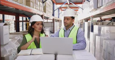 Güvenlik kaskı ve yansıtıcı yelek giyen iki depo çalışanı depolama tesisinde masada oturup, lojistik belgelerini ve dizüstü bilgisayardaki kutular dolusu verileri incelemeye odaklandılar.