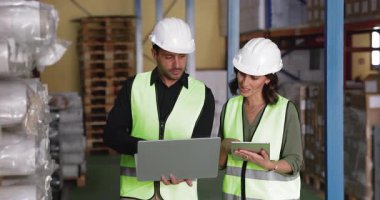 Latin erkek ve kadın iş arkadaşları depoda birlikte çalışıyorlar, sıkı şapkalar ve güvenlik yelekleri giyiyorlar, dizüstü bilgisayarlara bakıyorlar, tablet bilgilerini kontrol ediyorlar, envanter verilerini analiz ediyorlar ve yönetiyorlar.