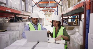 Koruyucu üniforma giyen iki depo çalışanı dizüstü bilgisayar kullanarak çalışıyor, hisse senedi bilgilerini kontrol ediyor ve güncelliyor, yeni siparişleri gözden geçiriyor, envanter yönetimiyle ilgileniyor, ürün miktarlarını teyit ediyor.