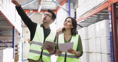 İki Hispanik dağıtım merkezi çalışanı raflara istiflenmiş karton kutularla dolu koridorda yürüyorlar, tablet kullanıyorlar, belgeleri gözden geçiriyorlar ve modern depoda birlikte çalışıyorlar.