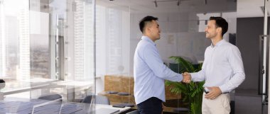 Modern ofiste el sıkışan iki çok ırklı işadamı şehrin ufuk çizgisine bakan geniş pencereli, yatay bir fotoğrafla. Karşılıklı saygı, işbirliğinin başlangıcı, anlaşma veya anlaşma kapatma