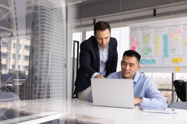 Danışman, toplantı veya modern ofiste brifing sırasında Asyalı erkek çırak için çevrimiçi görev tamamlamada yardımcı olur, dizüstü bilgisayara bakın, ekranda gösterilen bilgileri gözden geçirin, sonuçları tartışın, verileri analiz edin