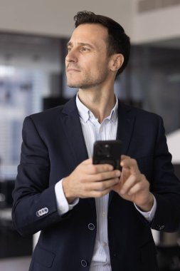 Düşünceli orta yaşlı iş adamı zarif resmi bir ceket giyer modern akıllı telefonu tutar çalışma yerinde tek başına durur, cevabı düşünür, e-posta ya da tebliğ üzerine düşünür, önemli bir iş aramasını bekler.