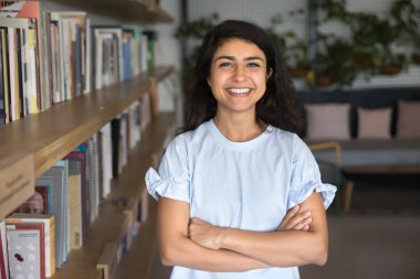 Genç, neşeli Hintli kadın kütüphanede ya da çalışma alanında duruyor çeşitli kitaplarla dolu raflarda duruyor. Çalışkan öğrenci, araştırmacı, yaratıcı profesyonel