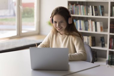 Uzaktan öğrenme ile meşgul. Uzakta odaklanmış genç kız öğrenci öğrenci çalışması. Öğretmenle iletişim kurmak için internet kamerası kullan. Kablosuz kulaklıklarda canlı sanal sınıfı dinle.