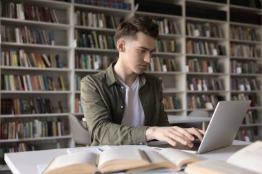 Uzaktan öğrenme. Meşgul genç üniversite öğrencisi kütüphane masasında çalışıyor ödev ödevi için kağıt bilimsel edebiyat el yazısıyla yazılmış notlar kullanarak dizüstü bilgisayar ödevi yazıyor.