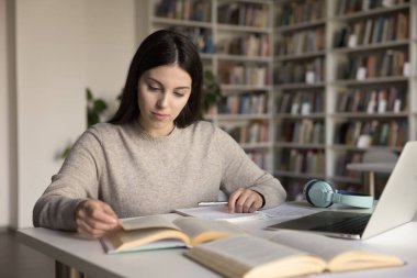 Yazmadan önce araştır. Kütüphane masasında kendi kendini eğiten meşgul bir kız öğrenci. Kağıt kitap oku. Yazılı ödevi desteklemek için dizüstü bilgisayar kullan.