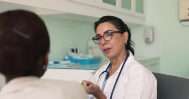 Ciddi, gözlüklü kıdemli İspanyol doktor muayene odasında Afrikalı hasta kadınla konuşuyor, danışmanlık yapıyor, tedavi önerisinde bulunuyor, tıbbi terapi görüyor.