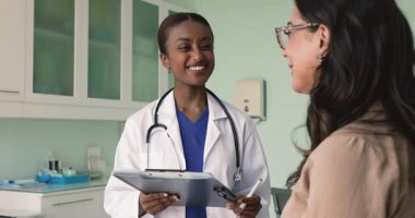 Olumlu genç Afrikalı doktor hasta kayıtlarını dolduruyor, toplantı formlarını dolduruyor, yaşlı kadınla dostça gülümsüyor, sağlık şikayetleri, terapi, tedavi, notlar hakkında sorular soruyor.