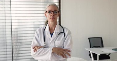 Muayene odasında güvenle poz veren yaşlı kadın doktor, kameraya gülümseyerek bakıyor, etrafa profesyonellik, deneyim ve güven saçıyor. Sağlık uzmanı, klinik ofis portresi