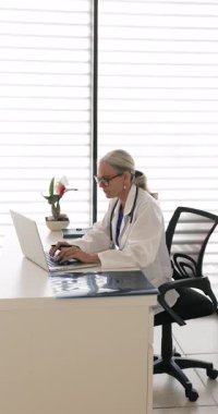 Beyaz önlüklü olgun kadın doktor iş yerinde dizüstü bilgisayarla oturur, dijital hasta kayıtlarını inceler, veri girer veya tanı notlarını günceller, tedavi planı yazar. Modern teknoloji kullanılarak çalışma akışı