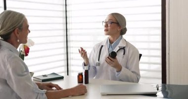 Kadın hekim danışma odasında oturuyor, ilaç şişesini tutuyor, olgun kadın hastaya ilaç dozunu açıklıyor, yeni ilaçlarını yazıyor, vitamin kompleksi öneriyor. Eczacılık.