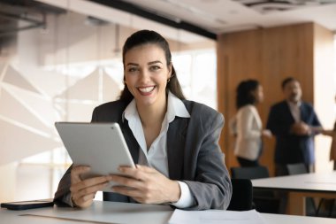 Profesyonel güven ve dijital aletler. İş yerinde gülümseyen genç bir iş kadını danışmanı elinde tabletle masada oturmuş müşteriyle tanışmaya hazır bir şekilde kameraya bakıyor.