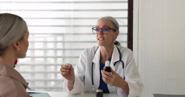 Beyaz önlüklü olgun kadın doktor muayene odasında oturuyor, ilaç şişesini tutuyor, kadın hastaya dozajı açıklıyor, iyi sağlık ve hastalık tedavisi için yeni ilaçlar veya vitamin kompleksi yazıyor.