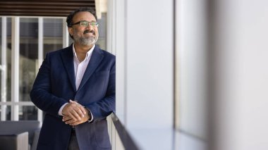 Ofiste pencereden bakan, gülümseyen, gayrimenkul yatırımını düşünen, finansal başarıyı düşünen neşeli, başarılı Hintli CEO. Kopya alanı ile günlük afiş portresi