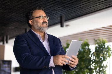 Positif kıdemli Hintli işadamı dijital tablet tutuyor, ofis salonunda modern internet teknolojisini kullanıyor, başka yere bakıyor, internet sektöründeki büyümeyi düşünüyor, çevrimiçi profesyonel iletişimi düşünüyor