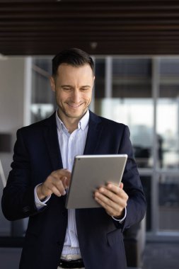 Mutlu genç Latin iş adamı tablet bilgisayarında yazı yazıyor, çevrimiçi ticaret hizmeti, finansal uygulama, iş için modern teknoloji, internette gezinme, gülümseme. Dikey çekim