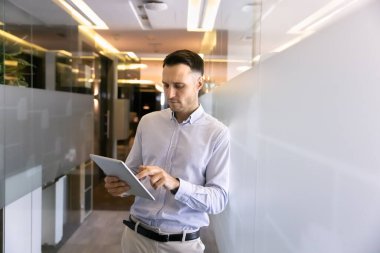 Ciddi anlamda başarılı İspanyol işadamı internet teknolojisini internet iletişimi için tablet olarak kullanıyor, ofis koridorunda duruyor, cam duvara yaslanıyor, dijital aygıtla yazı yazıyor..