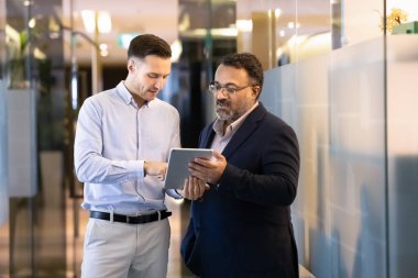 Ciddi genç iş yazılımı uzmanı Hintli kıdemli meslektaşına tablet için yeni başvuru formunda yardım ediyor. Çeşitli yaşlardaki meslektaşlar web servisini birlikte kontrol ediyorlar.