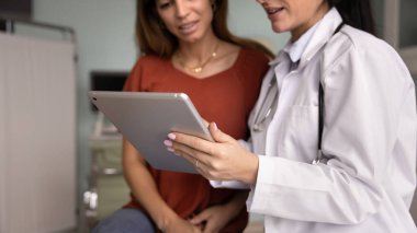 Kadın doktorun elleri tablet tutuyor, muayene için elektronik kayıtları kullanıyor, cihazdan hastaya muayene sonuçlarını gösteriyor, check-up sonuçlarını, reçeteyi açıklıyor. Kırpılmış afiş atışı