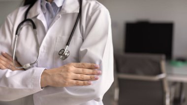 Doktor önlüğü ve steteskop giymiş, kollarını katlayıp güven jesti yaparak klinikte poz veren kadın sağlık görevlisinin kırpılmış görüntüsü. Tanımlanamayan bir tıbbi çalışanla kesilmiş afiş.