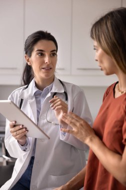 Ciddi genç İspanyol kadın pratisyen doktor muayeneden sonra kanepede oturan hastayla konuşuyor, kontrol sonuçlarını, tedaviyi, tedaviyi, reçeteyi, dijital tableti elinde tutmasını tartışıyor. Dikey çekim