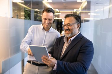 Genç ve yaşlıların çeşitli erkek proje arkadaşları tabletten komik içerik izliyorlar, ofis salonunda duruyorlar, iş yerinde alet kullanıyorlar, cihaza işaret ediyorlar, gülüyorlar.