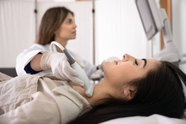 Genç hasta kadın ultrason tiroid kontrolü için kliniğe gidiyor, kanepede yatarken tıbbi teknisyen boyuna tarayıcı uyguluyor, lenf düğümlerini ve boğazını inceliyor. Yan görünümü kapat