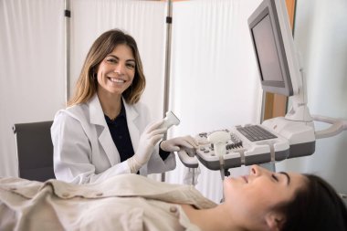 Pozitif ultrason teşhisi teknisyeni beyaz laboratuvar önlüğü giyen doktor sonografi tarama makinesinde çalışıyor ve hasta kanepede yatıyor, kameraya bakıyor, gülümsüyor.