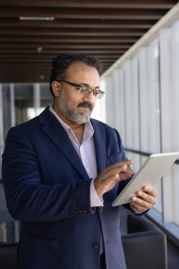 Modern teknolojiyi kullanan olgun Hintli işadamı, finans yönetimi için online uygulama, çevrimiçi ticaret, dijital tablet üzerinde daktilo, ofis salonunda durmak, internette gezinmek. Dikey çekim