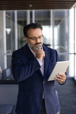 Düşünceli Hintli yaşlı CEO dijital tablet bilgisayardan online içeriği okuyor veya izliyor, çeneye dokunuyor, yatırım yönetimi hakkında düşünüyor, ticaret stratejisi, çevrimiçi teknoloji