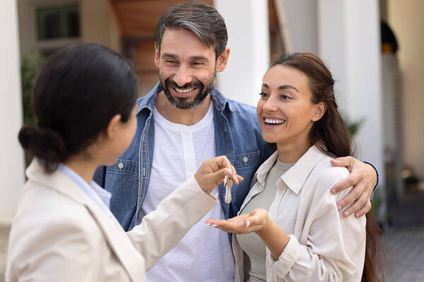 Married couple exuding joy of receiving keys to new house. Realtor handing over keys to spouses, standing together on porch of their first dwelling. Successful completion of real estate transaction