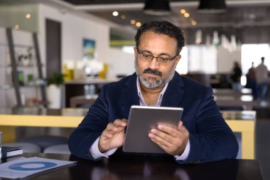 Ciddi Hintli yönetici açık ofis iş yerinde tablet üzerinde yazı yazıyor, online iletişim teknolojisini pazarlama analizi için kullanıyor, okuyor, içeriği izliyor.