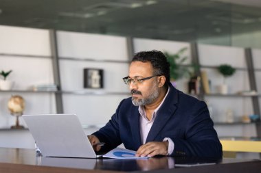 Ciddi meşgul yaşlı Hintli yönetici pazarlama analizi için online iş uygulaması kullanıyor, İnternet satış istatistiklerini kontrol ediyor, iletişim için dizüstü bilgisayarda yazıyor.