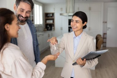 Emlakçı kadın neşeli Latin çiftin anahtarlarını teslim ediyor, şık mutfak iç dekorasyonu olan rahat bir ev odasında yan yana duruyorlar. Gayrimenkul işlemlerinin tamamlanması, mülkiyet veya kiralama