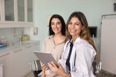 Mutlu, genç, Latin doktor kadın elinde tablet, pozitif kadın hasta bir kadın tarafından kanepeye oturmuş, dişlek gülümsemeyle kameraya bakıyor, sağlık hizmeti profesyonel portresi için poz veriyor.