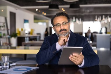 Düşünceli yaşlı Hintli iş adamı birlikte çalıştığı ofiste tabletle çalışıyor, çenesine dokunuyor, başka yere bakıyor, internet projesine yaratıcı bir çözüm düşünüyor.