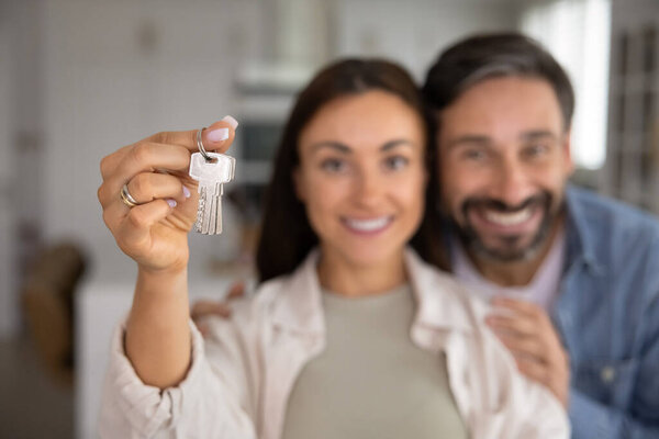 Крупный план руки с кучей ключей от дома на размытом фоне улыбающейся латинской супружеской пары, смотрящей в камеру с радостным выражением лица. Покупка дома через банковскую ипотеку, аренду, переезд