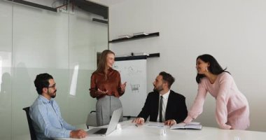 Çok ırklı iş grupları, takım çalışması için büyük bir masada toplanıyorlar, toplantı salonunda konuşuyorlar, proje fikirleri tartışıyorlar, gülümsüyorlar. Kadın lider, koç, proje yöneticisi meslektaşlarıyla konuşuyor, gülümsüyor.
