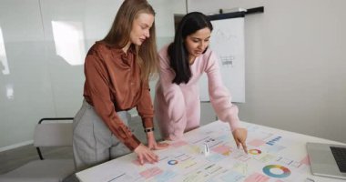 Toplantı masasındaki kağıt yığınlarını analiz eden, satış tablolarına işaret eden, analitik verileri, iş büyüme stratejisini tartışan çok ırklı genç kadın pazarlama uzmanları.