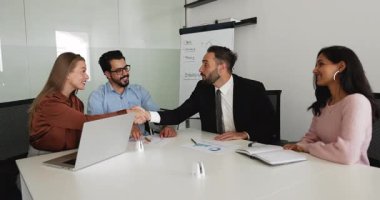 Çok ırklı genç iş ortakları ofis masasında buluşur, el sıkışır, selamlaşır, etkin ortaklık anlaşması yapar, pazarlığı başlatır veya bitirir, gülümser.
