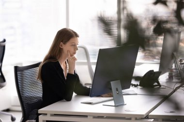 Ciddi genç 20 'li yaşlardaki finans uzmanı kadın açık ofis alanındaki bilgisayar monitöründe çalışıyor, çeneye dokunuyor, iş için online yaratıcı çözüm düşünüyor. Gizli çekim.