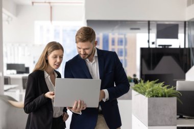 Kurumsal takım elbiseli iki genç iş arkadaşı birlikte dizüstü bilgisayardan içeriği izliyorlar, birbirlerine danışıyorlar, ellerinde aletlerle ofiste duruyorlar, konuşuyorlar, bilgisayar kullanarak takım çalışması yapıyorlar.