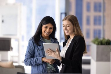 İki mutlu çok ırklı kadın iş arkadaşı dijital teknolojiyi takım çalışması için tablette kullanıyorlar. Ürün yöneticisi şirket internet proje sunumunu pozitif meslektaşına gösteriyor